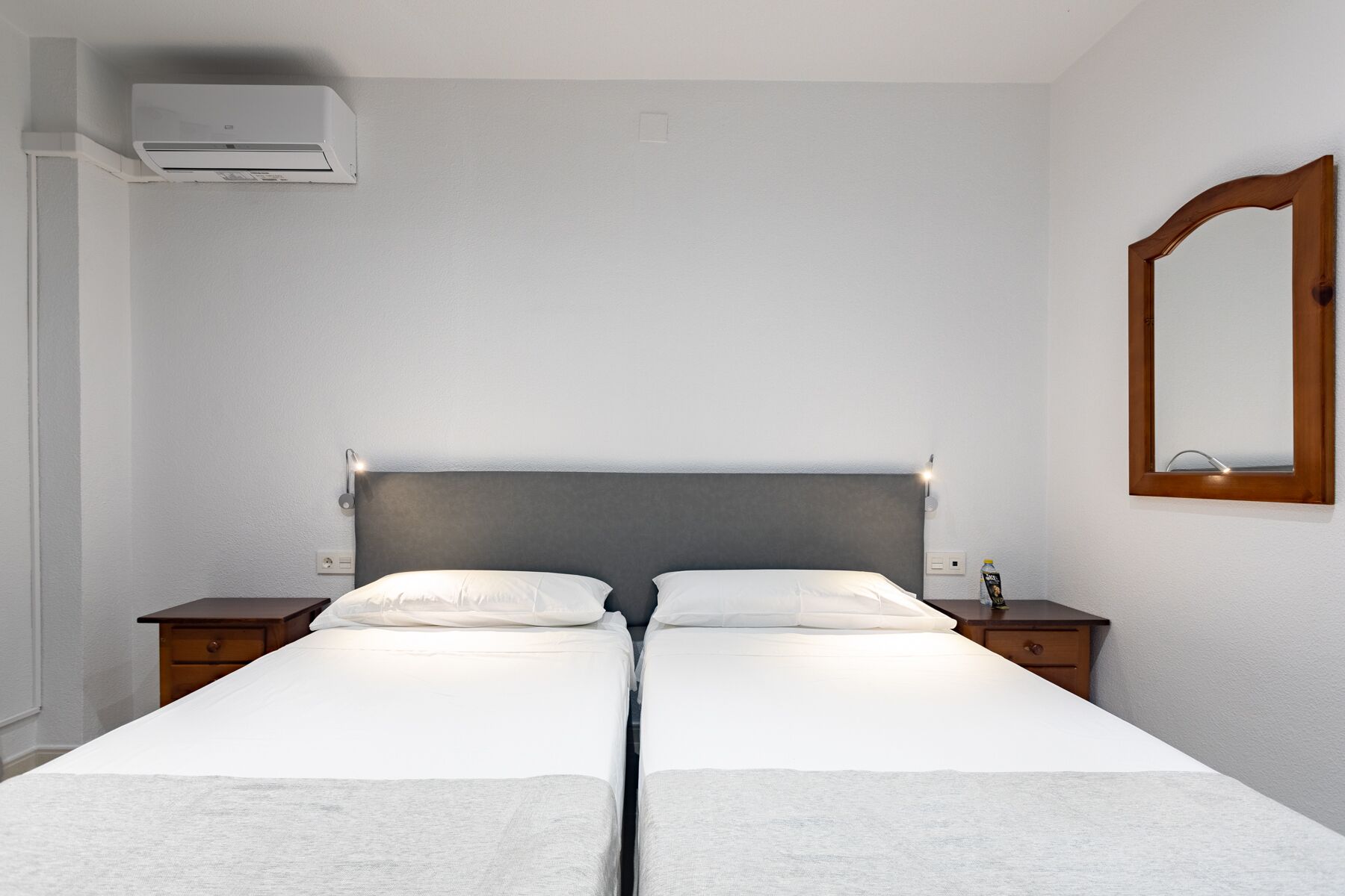 Una cama doble con cabecera gris, dos mesitas de noche, un espejo montado en la pared y un aire acondicionado instalado en el techo.