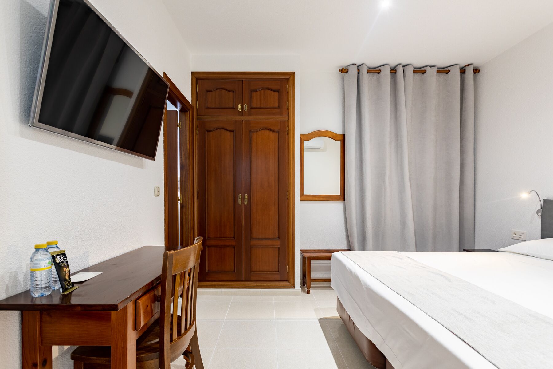 Un dormitorio con una cama, un escritorio con silla, un televisor montado en la pared, un armario y una ventana con cortinas.