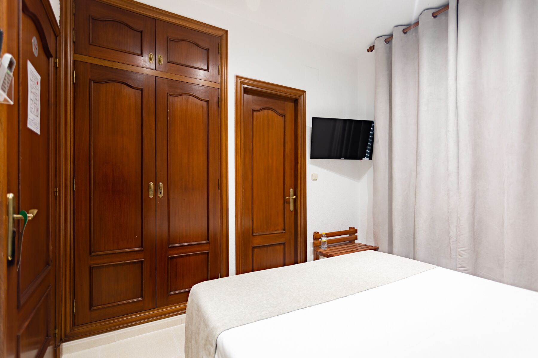A bedroom with a bed, wooden wardrobe, a television mounted on the wall, and a curtain.