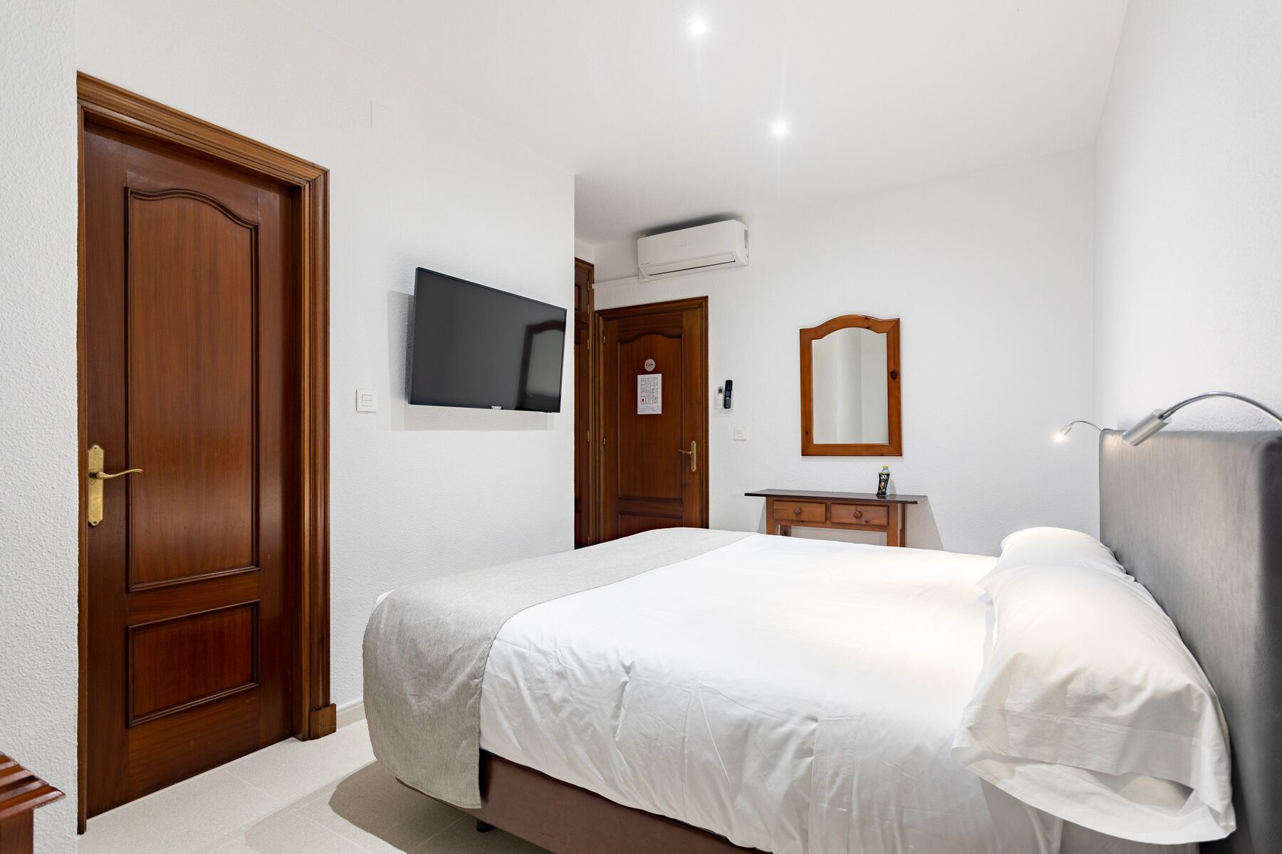 A hotel room with a bed, a TV, a mirror, and a desk with a lamp.