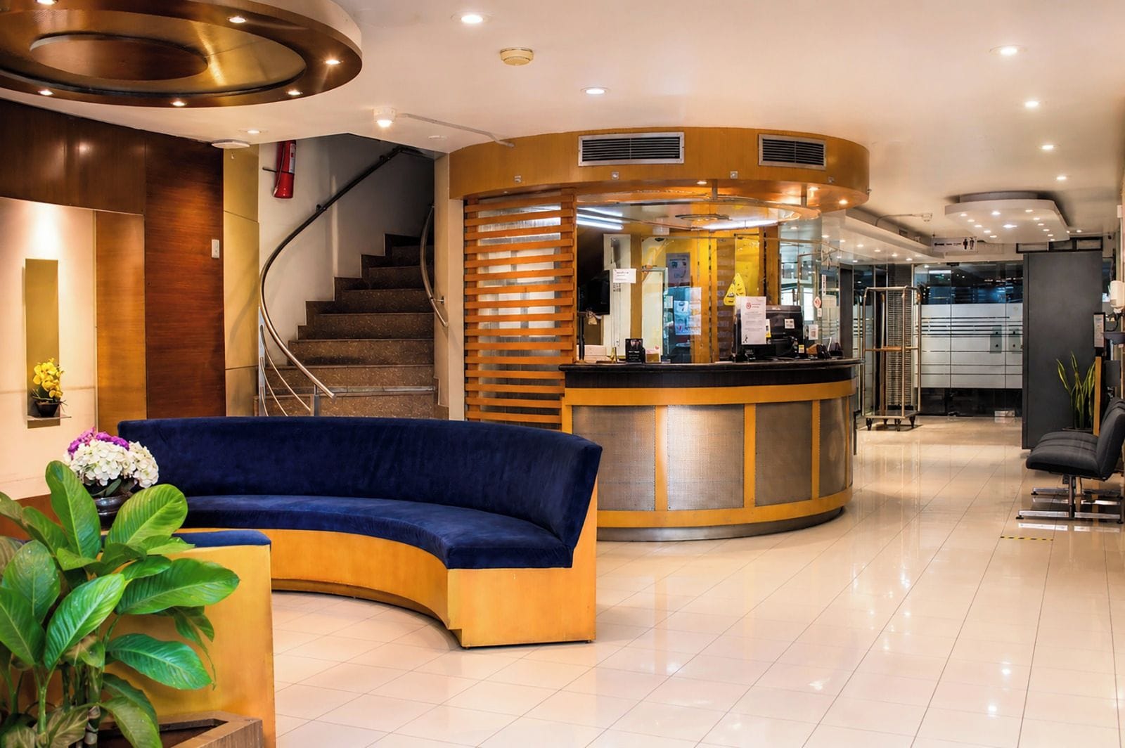 Un moderno vestíbulo de hotel con un mostrador de recepción curvo, un sofá azul, una escalera y una planta en maceta.
