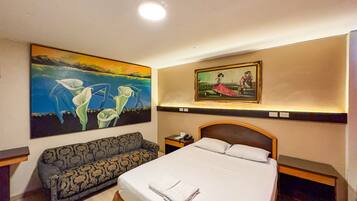 A hotel room with a bed, a sofa, a nightstand, and two paintings on the wall.