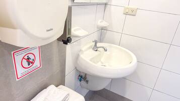 Un baño con un lavamanos blanco, un inodoro y un secamanos. Hay un cartel que recomienda no tirar papel higiénico.