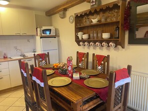 A dining area with a wooden table set for four, surrounded by chairs with red and white cloths. A microwave and a refrigerator are visible in the background. - La Hussaire - The Gingerbread House Cottage (Beauficel)
