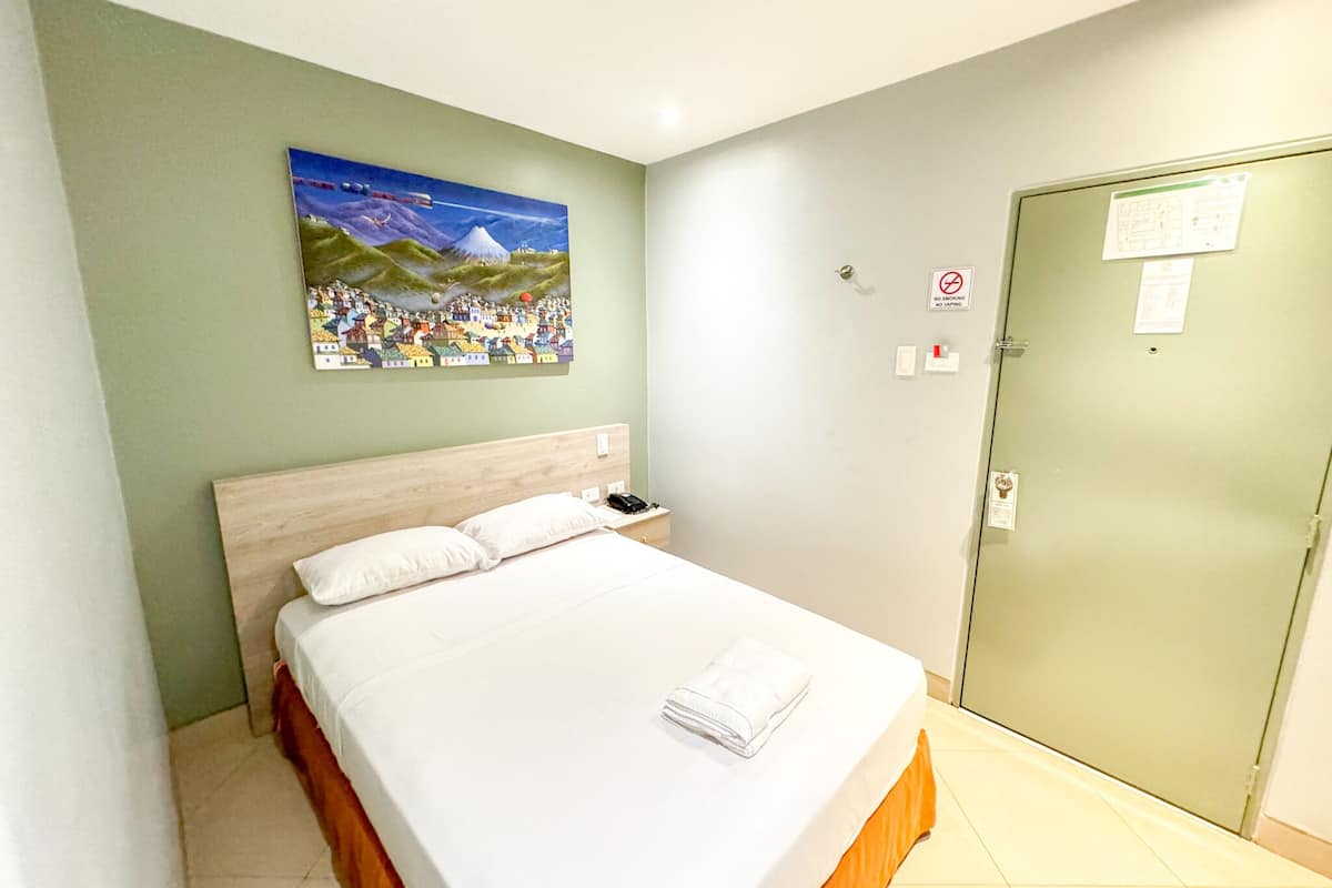 A hotel room with a bed, a painting on the wall, a door with a no smoking sign, and a light switch.