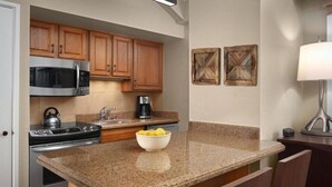 A compact kitchen with wooden cabinets, a microwave, stove, and a coffee maker. There is a small island with a bowl of lemons and a chair. Two wooden wall art pieces are visible. - Elegant Streamside at Vail Studio with Outdoor Pool (Vail)