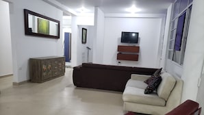 Una sala de estar con un sofá marrón, un sofá blanco, una silla roja, un aparador de madera con diseños florales, un televisor montado en la pared y un cuadro enmarcado sobre el televisor.