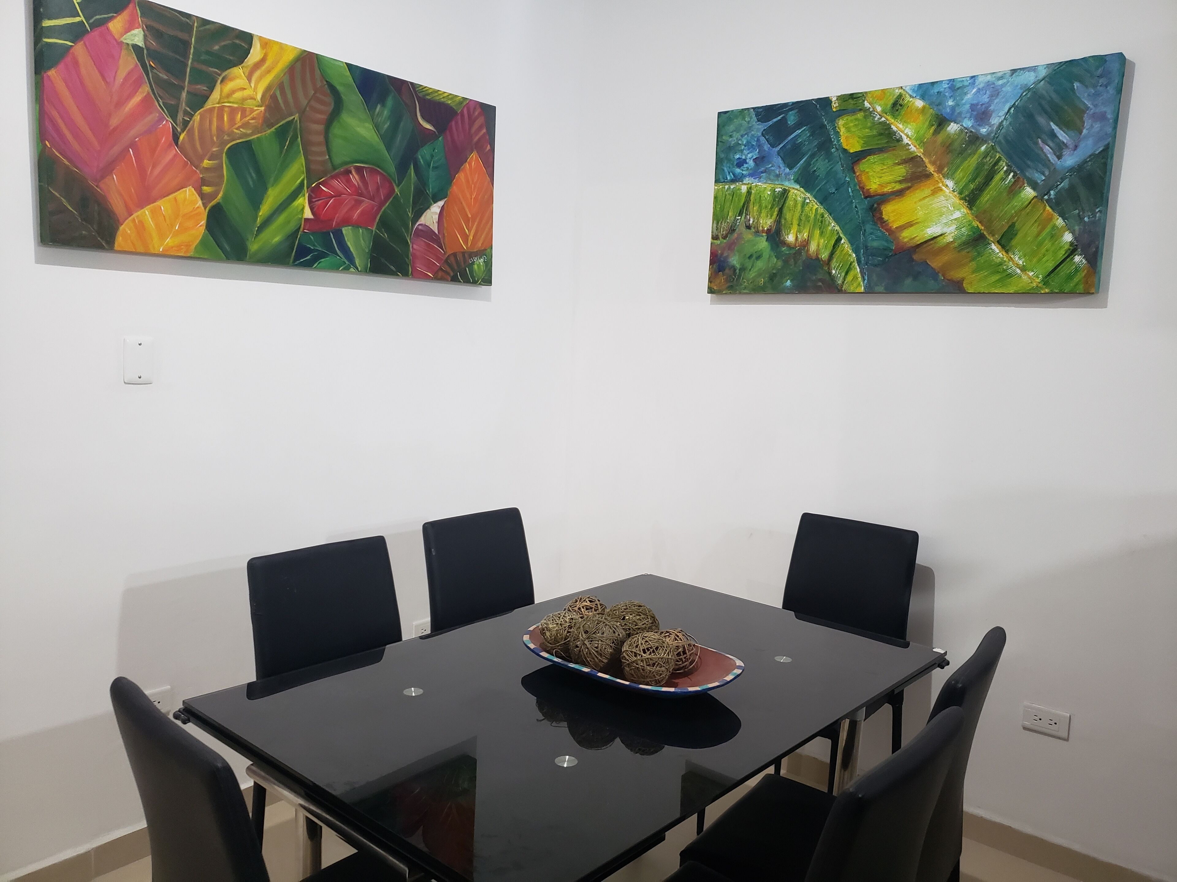 Uma sala de jantar com uma mesa e cadeiras pretas, uma tigela com bolas decorativas e duas pinturas coloridas na parede.