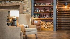 Un salon chaleureux avec une cheminée, un fauteuil confortable, une table d’appoint avec une lampe et une bibliothèque en bois remplie d’objets décoratifs.