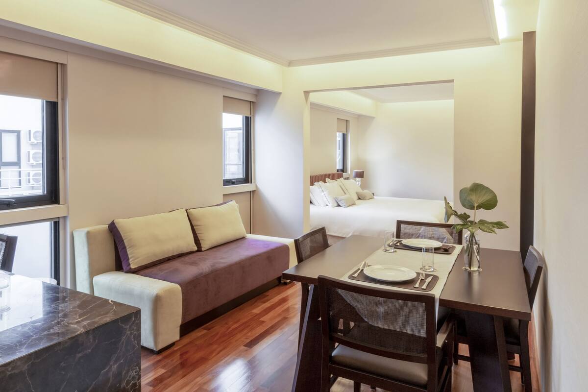 A modern hotel room with a dining area, a sofa, a bed, and a large window.