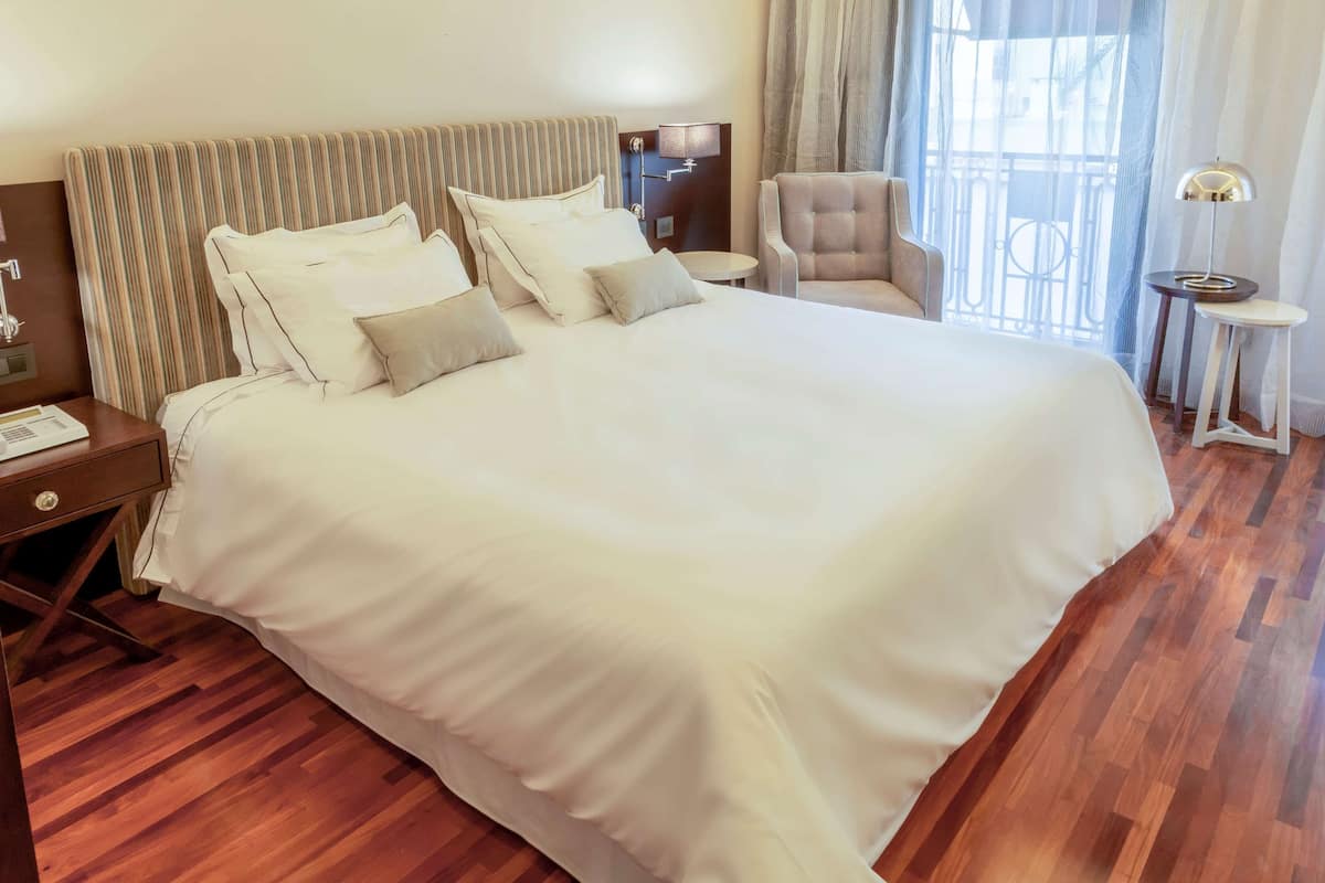 A hotel room with a large bed, a chair, a small table, and a window with curtains.