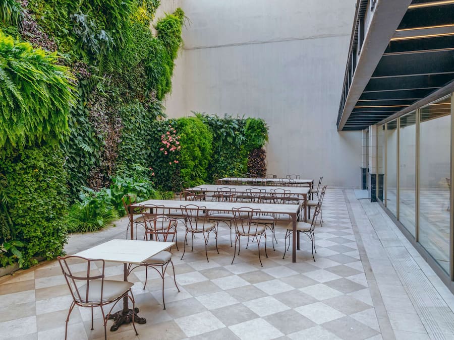 A courtyard with tables and chairs, surrounded by lush greenery and a checkered floor.