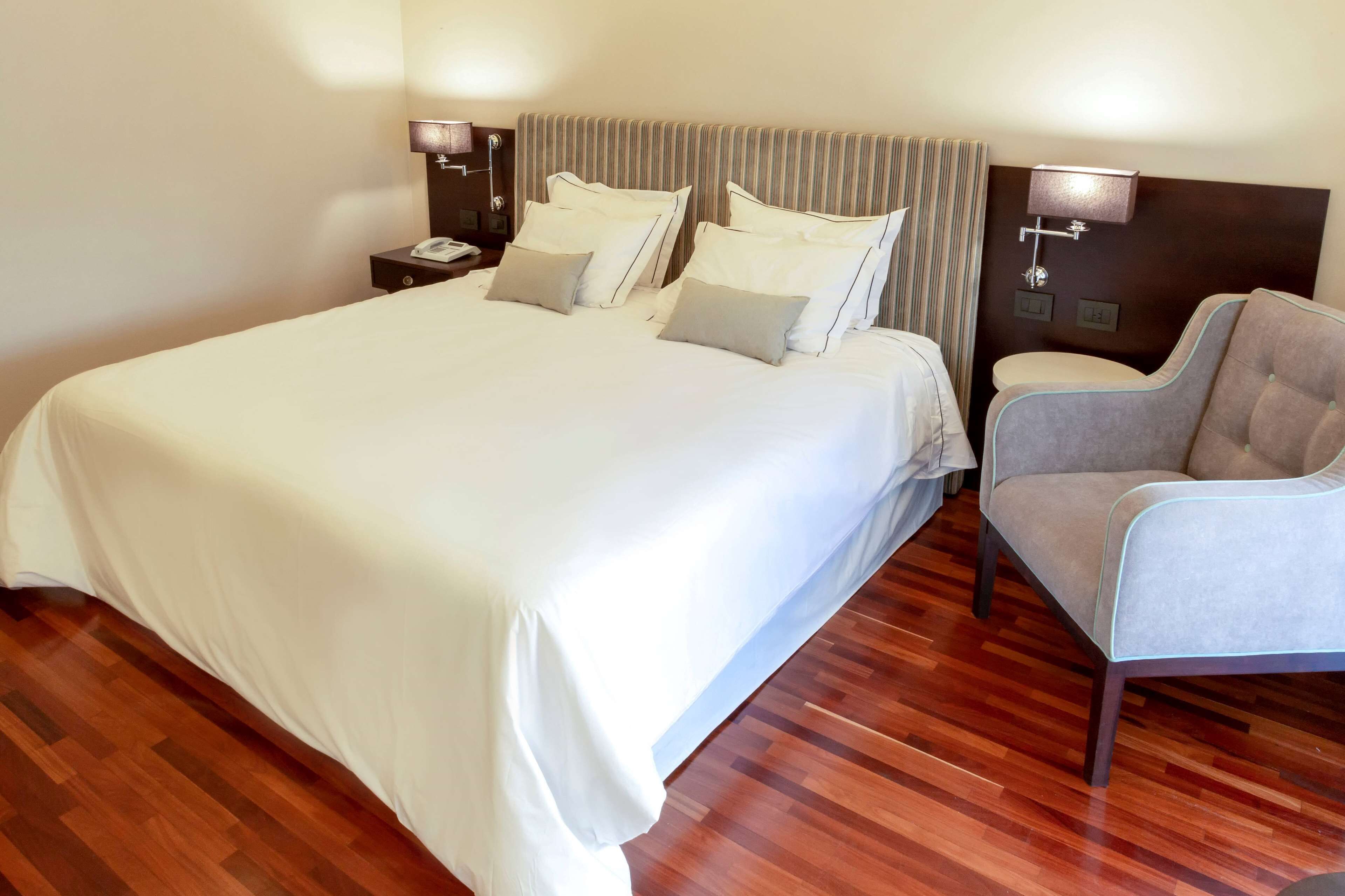 Una habitación de hotel con una cama grande, dos mesitas de noche con lámparas, una silla y suelo de madera.