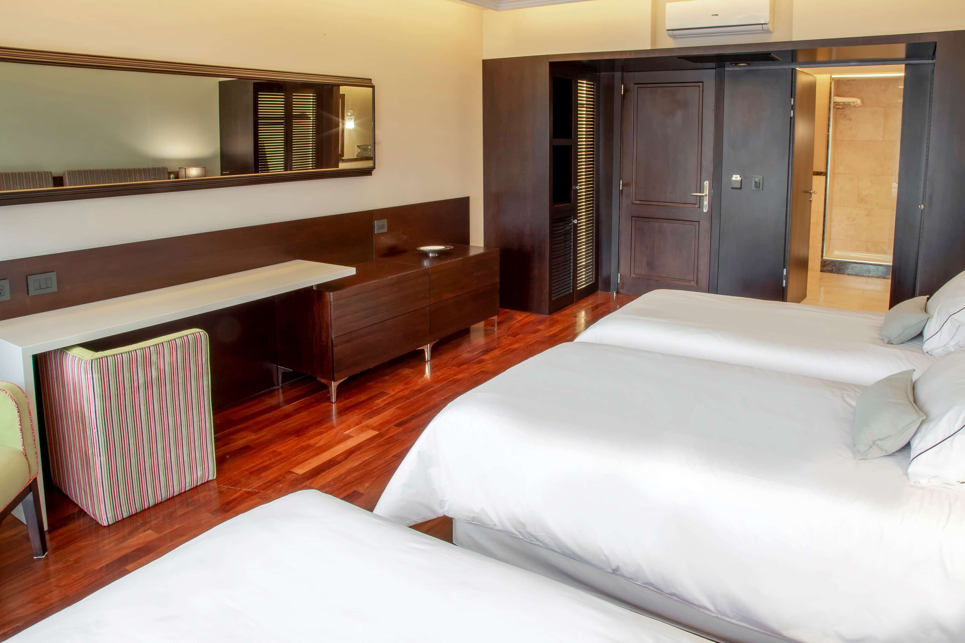 A hotel room with two beds, a vanity with a mirror, a wooden floor, and a wardrobe with a door leading to a bathroom.