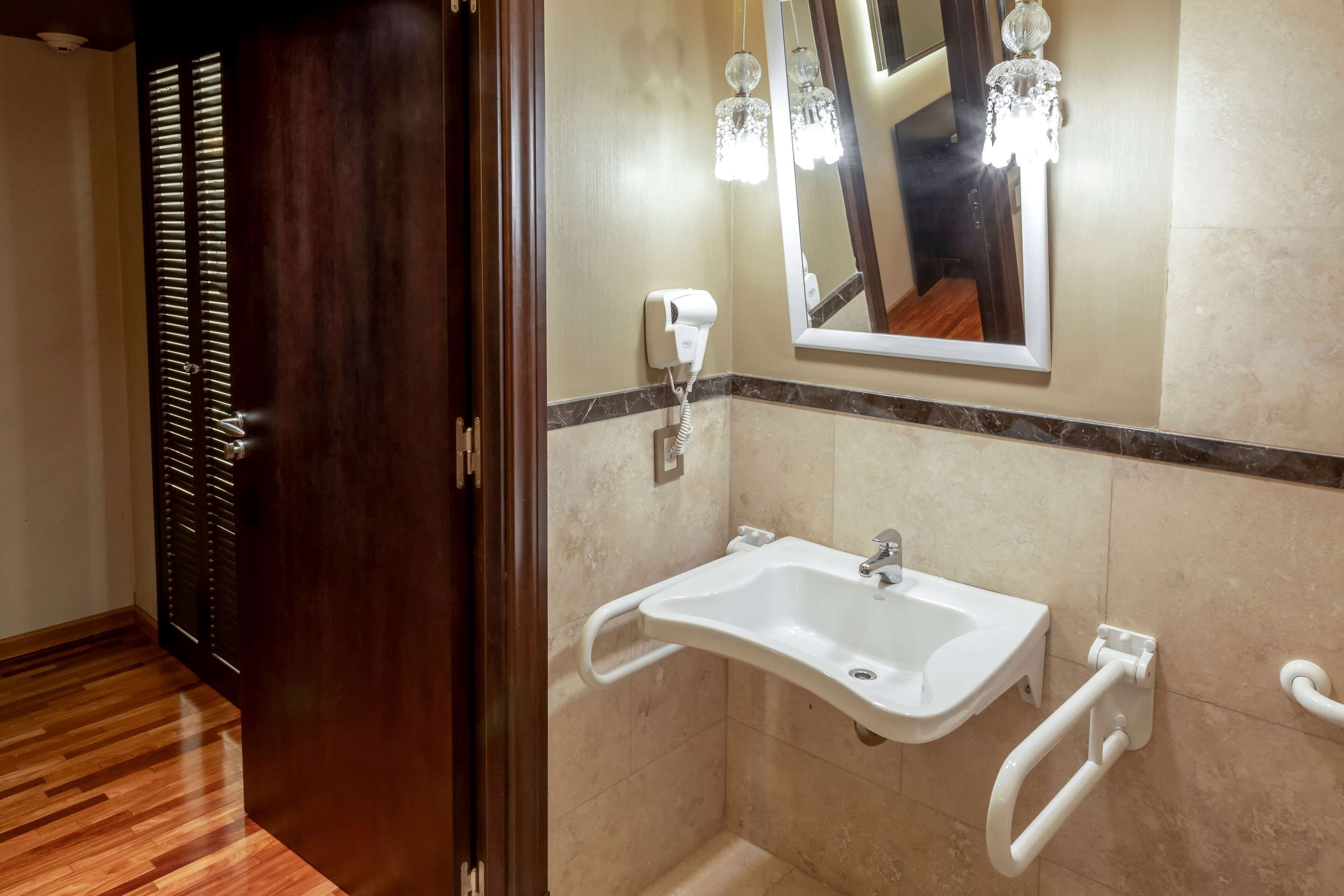 Une salle de bain avec un lavabo blanc, un miroir, un sèche-cheveux et une barre d’appui.