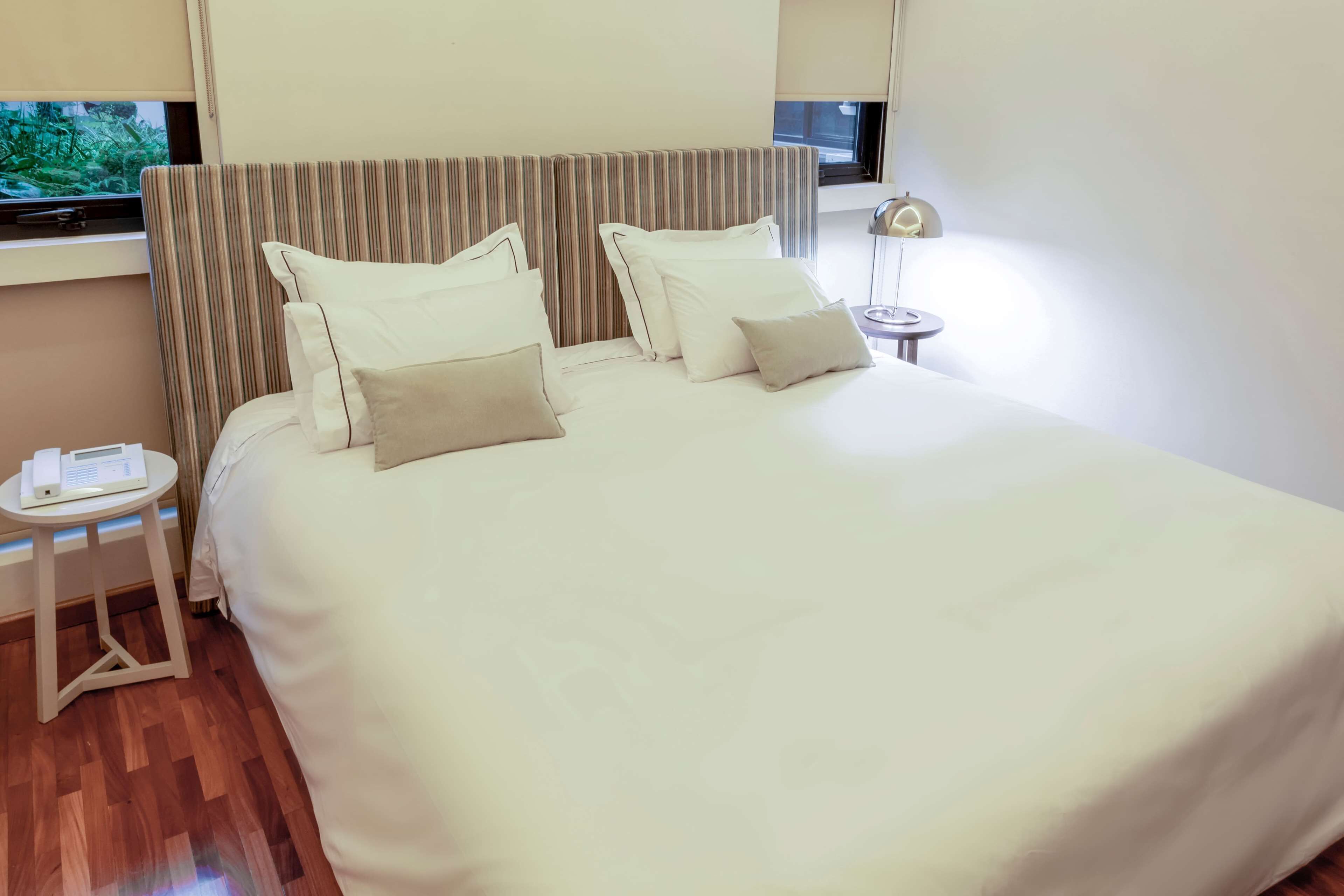 Una cama bien tendida con ropa de cama blanca y beige, un cabecero a rayas y una mesita de noche con una lámpara.