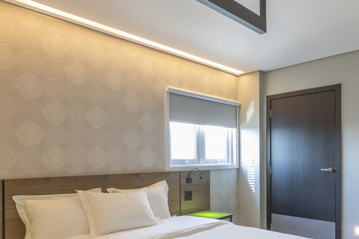 A modern hotel room with a large bed, a bedside table, a window with blinds, and a door.