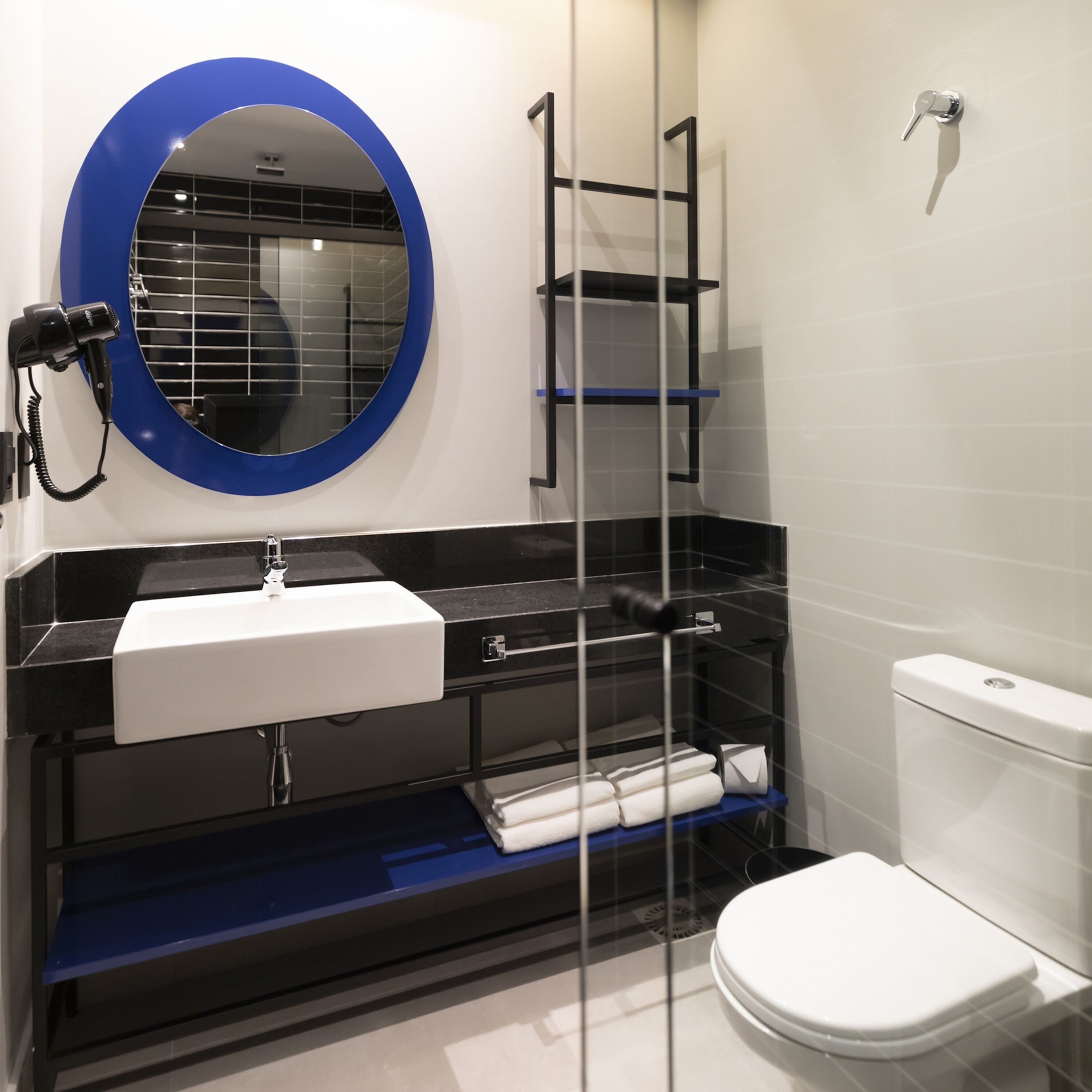 A modern bathroom with a white sink, black shelves, a round mirror, and a white toilet.