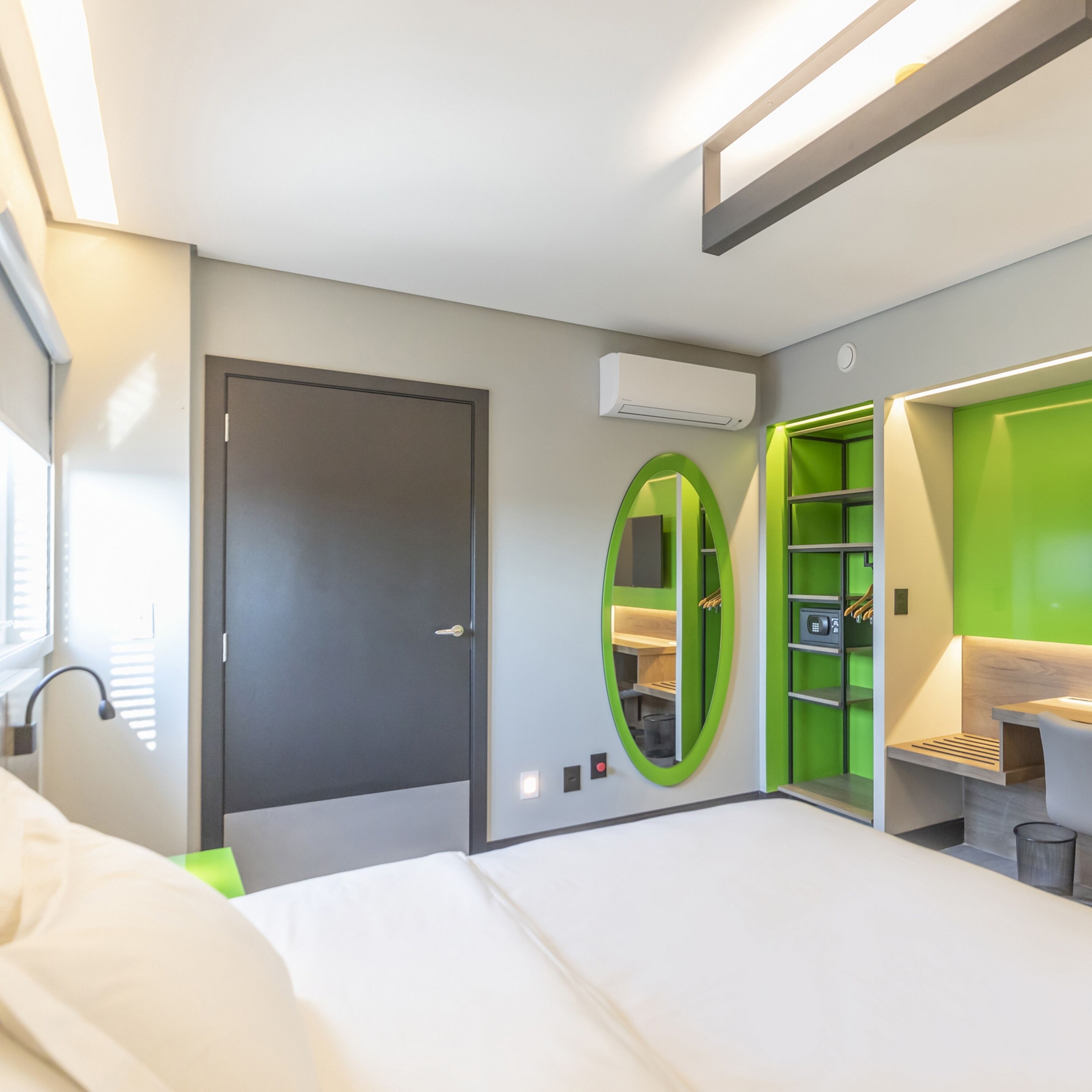 A modern hotel room with a bed, a desk with a chair, a green-framed mirror, and a wardrobe.