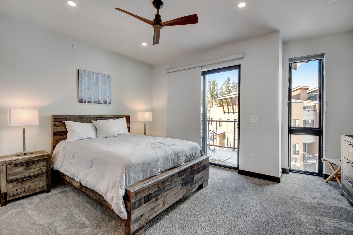 A bedroom with a wooden bed, two bedside tables, a ceiling fan, a large window, and a view of the outside.