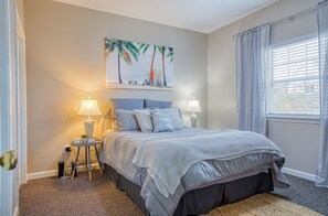 Une chambre à coucher avec un lit, deux tables de chevet avec des lampes, un tableau représentant une scène de plage accroché au mur, et une fenêtre avec des rideaux.