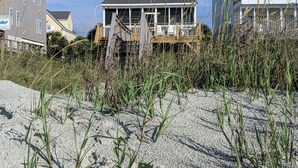 Ett tvåvåningshus med en balkong, omgivet av sand och strandväxtlighet.