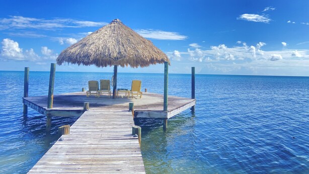 Een houten pier die leidt naar een gebouwtje met een rieten dak en twee stoelen.
