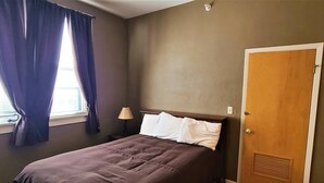 A bedroom with a bed, a nightstand, a lamp, a window with curtains, and a door. - The Westchester 1 BR located On The Bricks (Fairbury)
