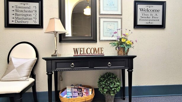 A reception desk with a 'WELCOME' sign, framed directional signs, a lamp, a chair, a basket of magazines, and a potted plant. - The Westchester 1 BR located On The Bricks (Fairbury)