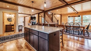 Une cuisine spacieuse dotée d’un grand îlot central, de poutres apparentes en bois au plafond et d’un coin repas avec une table et des chaises.