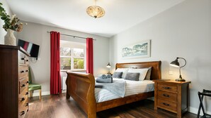 Ein Schlafzimmer mit einem hölzernen Bett, einem Schminktisch, einem Schreibtisch, einem Stuhl, einem Fernseher und einem Fenster mit roten Vorhängen.