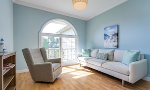 A living room with a light blue wall, a white sofa with patterned cushions, a grey armchair, a wooden floor, and a large window with a view of greenery.