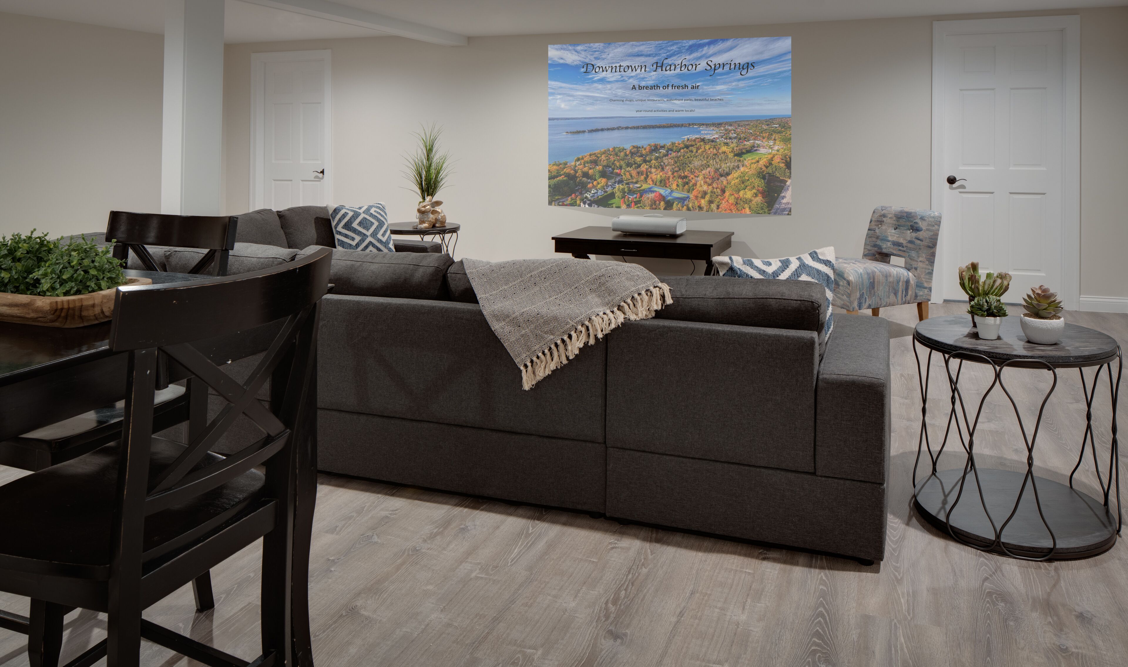 A modern living room with a grey sofa, a dining table, and chairs. There is a large poster on the wall and a small round table with plants.