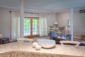 A living room with a large window, a dining area with a table and chairs, and a kitchen counter with a bowl and decorative items.
