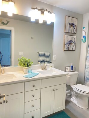 A bathroom with a white sink, a mirror, and a toilet. There are three framed pictures on the wall, a potted plant, and a blue towel on the counter.