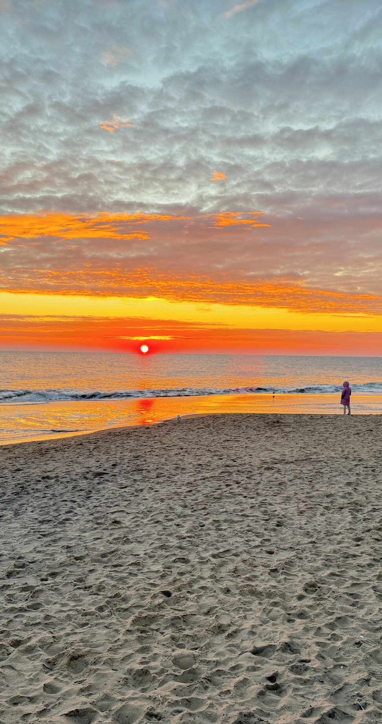 Una playa al atardecer, con olas y una persona de pie sobre la arena.