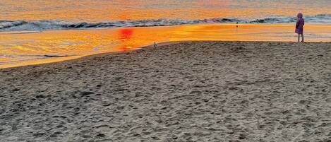 Una playa al atardecer, con olas y una persona de pie sobre la arena.