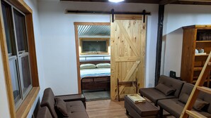 Uma sala de estar acolhedora com um sofá, um banquinho e uma escada de madeira. Uma porta de madeira com design de trave leva a um quarto com uma cama e uma janela.