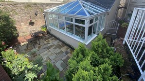 A conservatory with a glass roof, surrounded by a garden with various plants and a patio area.