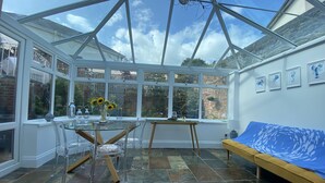 A conservatory with a glass roof, a dining table with chairs, a sofa, and a view of the garden through large windows.