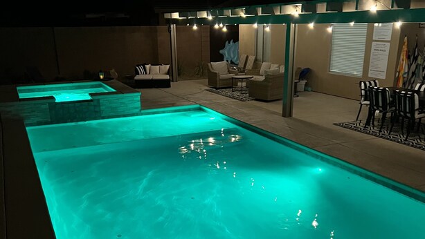 Una piscina con luz verde azulada, rodeada de asientos exteriores y una zona cubierta con guirnaldas de luces.