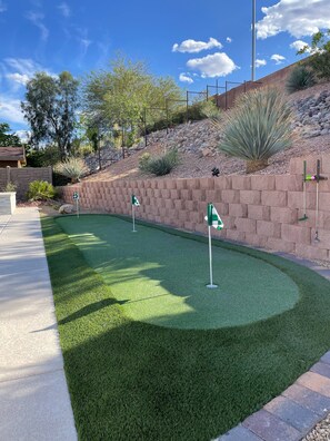 A well-maintained artificial green turf golf course with multiple holes, surrounded by a landscaped area with rocks and desert plants.