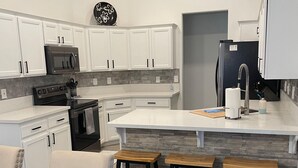 Una cocina moderna con armarios blancos, un microondas negro, una estufa y un refrigerador. Hay tres taburetes de madera y una mesa.