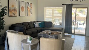 Una sala moderna con un sofá modular, dos sillones, una mesa de centro y un ventilador de techo. Hay tres cuadros en la pared, una ventana con persianas y una puerta corrediza de vidrio que da al exterior.
