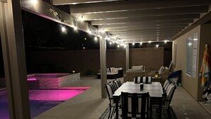 Un patio al aire libre con piscina, una mesa de comedor con sillas y luces colgantes.
