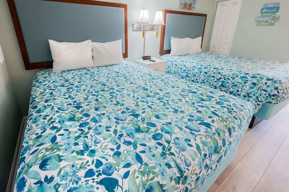 Une chambre d’hôtel avec deux lits, chacun recouvert d’une couvre-lit à motifs floraux, une table de chevet avec une lampe et un élément décoratif mural.