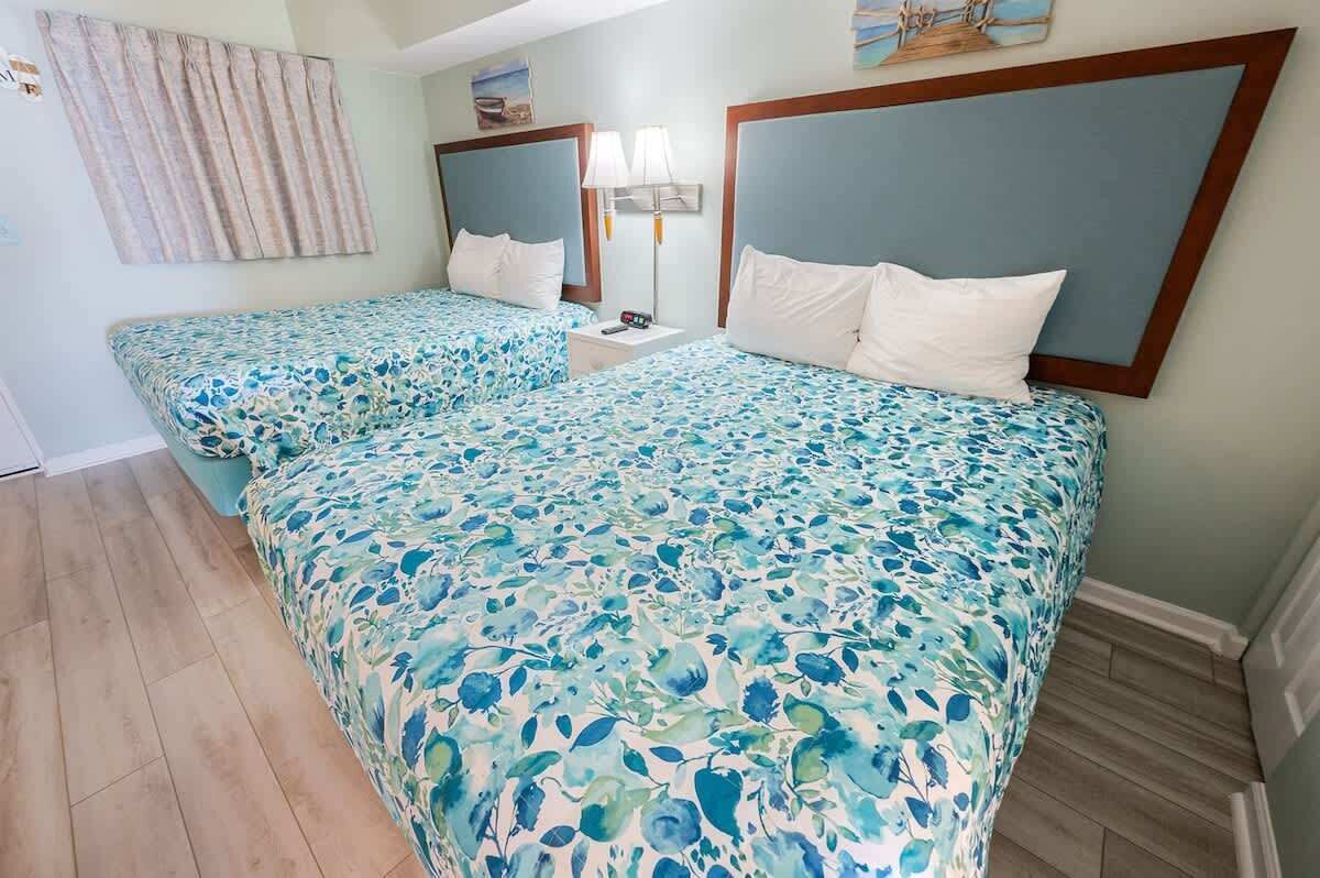 A hotel room with two beds, each with a floral bedspread, a nightstand with a lamp, and a painting on the wall.