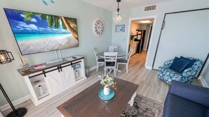 A living room with a wooden coffee table, a TV mounted on the wall, a dining area with white chairs, and a blue sofa. - Bay Watch 503 Direct Oceanfront w/ Tiki Bar, Pools (North Myrtle Beach)