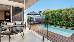 A modern outdoor pool area with a wooden deck, a seating area with a sofa and chairs, and a glass wall providing a view of the surrounding greenery. - Enjoy the deluxe swimming pool surrounded by a poolside sundeck (Lennox Head)