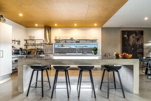 A modern kitchen with a central island, bar stools, and a large window. - Enjoy the deluxe swimming pool surrounded by a poolside sundeck (Lennox Head)